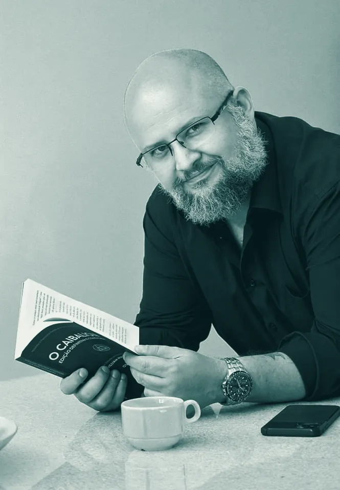 Gogo Cristo Neto sorrindo, com um livro aberto nas mãos e uma xícara de café sobre a mesa